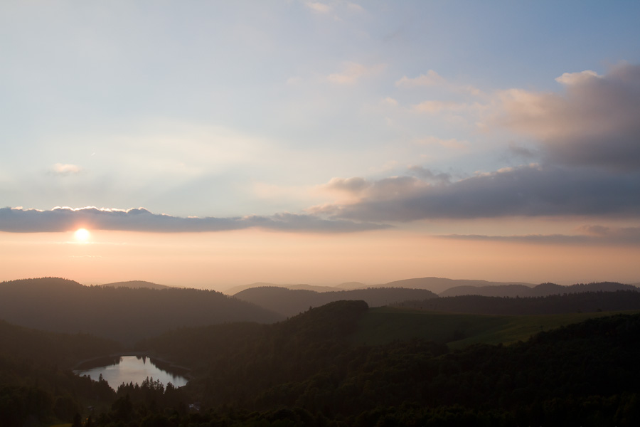 Soleil couchant sur le lac des Landes vu de loin entouré de l'ombre des sapins.
